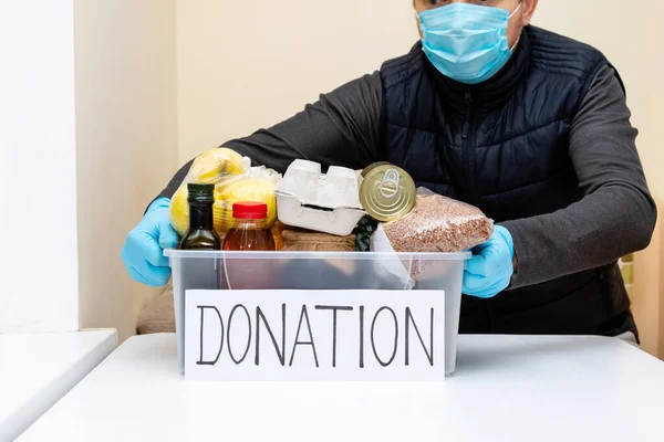Man volunteer in protective medical mask and gloves puts products in donation box. Work in fund charity food. Help, donate during quarantine coronavirus.