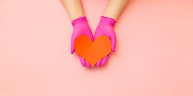 Kırmızı koruyucu eldivenli kadın eller pembe arka planda bir kalp sembolü tutuyor. Konsept - tıp, kardiyoloji. Boşluğu kopyala.