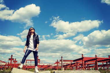 Deri ceketli ve güneş gözlüklü bir kadın tamamlanmamış bir binanın arka planında. Terk edilmiş yerlere git.