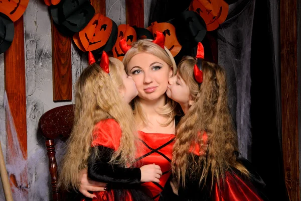 halloween scene with three attractive witches, young mother and two little daughters twins, in costumes in black and red indoors, sisters kissing their mommy