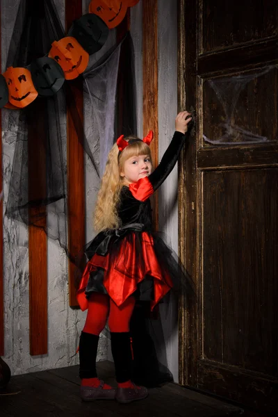 可爱的万圣节魔鬼女孩与金发与牛角在服装在红色和黑色打开旧木门室内 — 图库照片