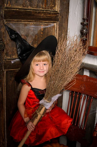 可爱的金发女郎万圣节女巫红色礼服和黑色帽子在旧木门室内扫帚 — 图库照片