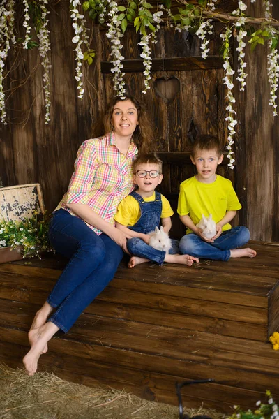 Mom and sons are sitting on wooden porch, holding two white fluffy rabbits in their hands, wisteria is blooming around