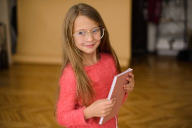 Okul çağında uzun saçlı, pembe kazaklı, gözlüklü, elinde not defteri olan güzel bir kız.