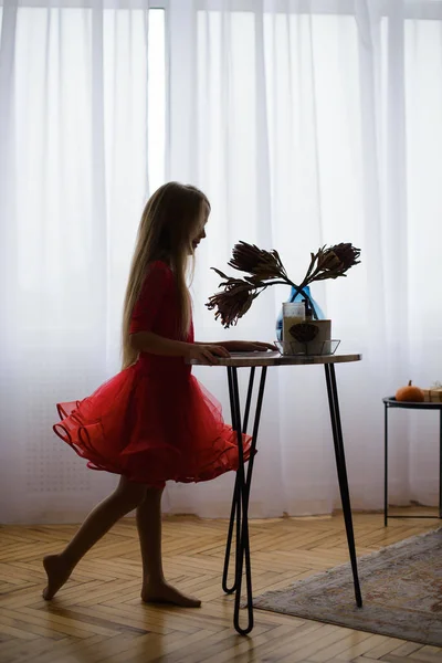 穿着红色舞衣的女孩的轮廓 在阳光下的窗前 在有木制花束的明亮房间里 站在靠近桌子的剖面上 开着花 — 图库照片