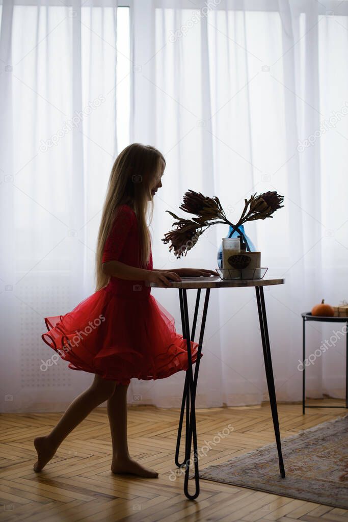 silhouette of girl in red dance dress, against background of window with daylight, in bright room of house with wooden parquet. Standing in profile near table with flowers