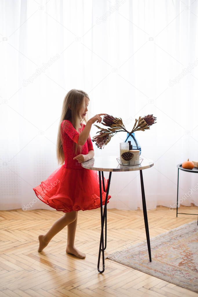 silhouette of girl in red dance dress, against background of window with daylight, in bright room of house with wooden parquet. Standing in profile near table with flowers