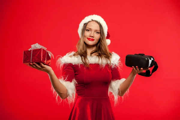 Natal, x-mas, inverno, conceito de felicidade - Linda jovem morena com cabelos longos em chapéu de Papai Noel usando fone de ouvido realidade virtual sobre fundo vermelho caixa de presente — Fotografia de Stock