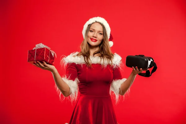 Natal, x-mas, inverno, conceito de felicidade - Linda jovem morena com cabelos longos em chapéu de Papai Noel usando fone de ouvido realidade virtual sobre fundo vermelho caixa de presente — Fotografia de Stock