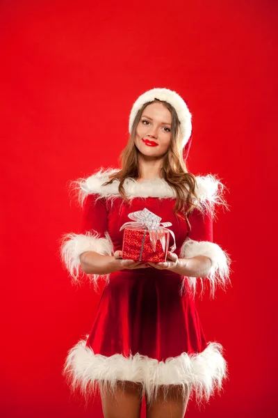 Karácsony, karácsonyi, téli, boldogság koncepció - mosolygó nő santa kalap segítő ajándék doboz, piros háttér felett — Stock Fotó