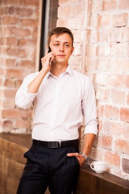Beyaz gömlekli yakışıklı işadamı tuğla duvarın yanında telefonda konuşuyor. Bir fincan kahve olduğunu
