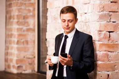 Siyah elbiseli genç yakışıklı işadamı kahve içme ve telefonda yakınındaki duvara sörf