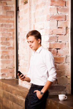 Beyaz gömlekli yakışıklı işadamı tuğla duvarın yanında telefonda konuşuyor. Bir fincan kahve olduğunu