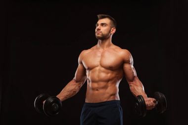 Spor salonunda yakışıklı atletik erkek bir spor salonunda dumbbells ile kasları yukarı pompalıyor. Fitness kas vücut üzerinde koyu arka plan izole.