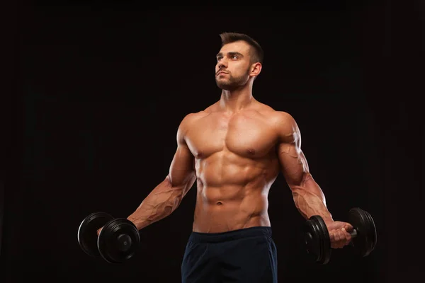 Bonito atlético na academia está bombeando músculos com halteres em um ginásio. Fitness corpo muscular isolado no fundo escuro . — Fotografia de Stock