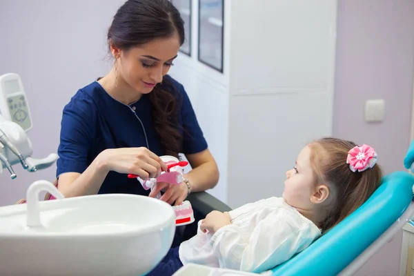 Pediatric dentist educating a smiling little girl about proper tooth-brushing, demonstrating on a model. Early prevention, raising awareness, oral hygiene demonstration concept. — Stock Photo, Image