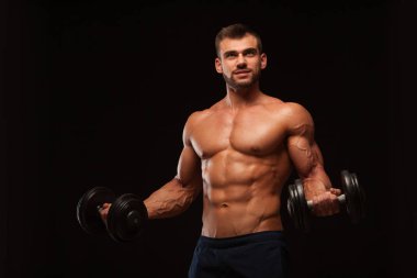 Spor salonunda yakışıklı atletik erkek bir spor salonunda dumbbells ile kasları yukarı pompalıyor. Fitness kas vücut üzerinde koyu arka plan izole.