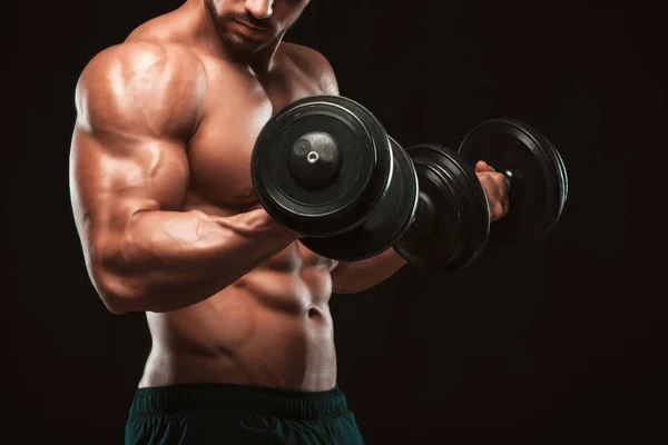 Hombre atlético de poder guapo en el entrenamiento bombeando músculos con pesas en un gimnasio. Fitness cuerpo muscular aislado sobre fondo oscuro. —  Fotos de Stock