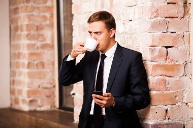 Siyah elbiseli genç yakışıklı işadamı kahve içme ve telefonda yakınındaki duvara sörf