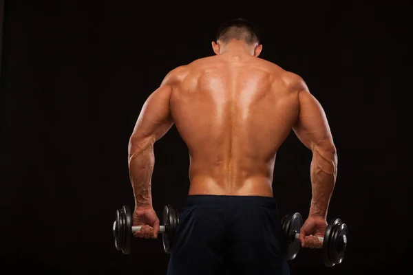 Muscular male model bodybuilder doing exercises with dumbbells, turned back. Isolated on black background with copyspace