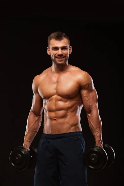 Spor salonunda yakışıklı atletik erkek bir spor salonunda dumbbells ile kasları yukarı pompalıyor. Fitness kas vücut üzerinde koyu arka plan izole. — Stok fotoğraf