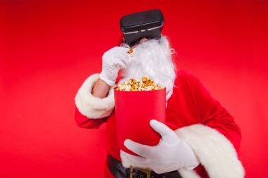 Santa Claus wearing virtual reality goggles and a red bucket with popcorn, on a red background. Christmas clipart