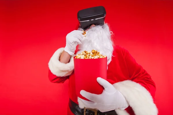 Babbo Natale indossa occhiali di realtà virtuale e un secchio rosso con popcorn, su uno sfondo rosso. Natale! — Foto Stock