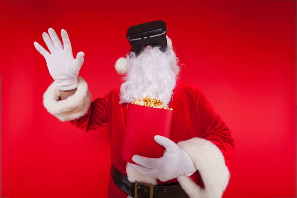 Santa Claus vistiendo gafas de realidad virtual y un cubo rojo con palomitas de maíz, sobre un fondo rojo. Navidad — Foto de Stock