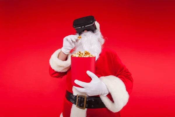 Santa Claus vistiendo gafas de realidad virtual y un cubo rojo con palomitas de maíz, sobre un fondo rojo. Navidad — Foto de Stock