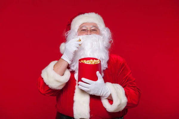 Tradicional Santa Claus viendo la televisión, comiendo palomitas de maíz. Navidad. Fondo rojo. emociones miedo sorpresa —  Fotos de Stock
