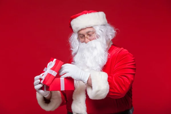 Weihnachten. photo santa claus macht Weihnachtsgeschenk und blickt in die Kamera, auf rotem Hintergrund — Stockfoto