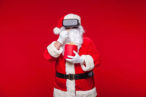 Babbo Natale indossa occhiali di realtà virtuale e un secchio rosso con popcorn, su uno sfondo rosso. Natale! — Foto Stock
