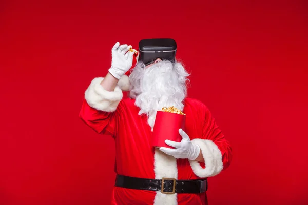 Weihnachtsmann mit Virtual-Reality-Brille und rotem Eimer mit Popcorn auf rotem Hintergrund. Weihnachten — Stockfoto