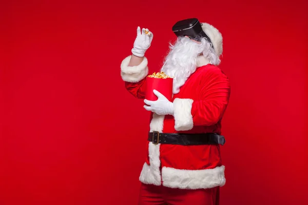 Santa Claus vistiendo gafas de realidad virtual y un cubo rojo con palomitas de maíz, sobre un fondo rojo. Navidad —  Fotos de Stock