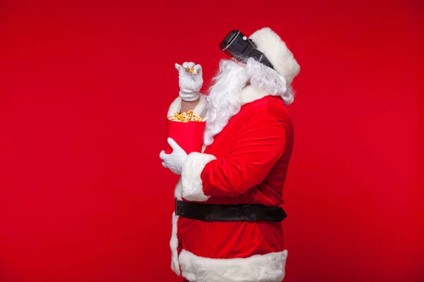 Santa Claus vistiendo gafas de realidad virtual y un cubo rojo con palomitas de maíz, sobre un fondo rojo. Navidad —  Fotos de Stock