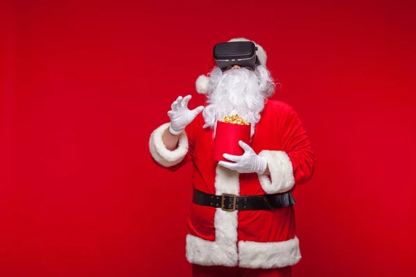 Santa Claus vistiendo gafas de realidad virtual y un cubo rojo con palomitas de maíz, sobre un fondo rojo. Navidad —  Fotos de Stock