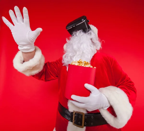Santa Claus vistiendo gafas de realidad virtual y un cubo rojo con palomitas de maíz, sobre un fondo rojo. Navidad — Foto de Stock