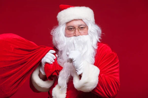 Retrato de Santa Claus con enorme saco rojo manteniendo el dedo índice por la boca y mirando a la cámara —  Fotos de Stock