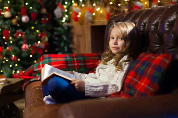 Happy little girl reading a story book by on the couch in a cozy