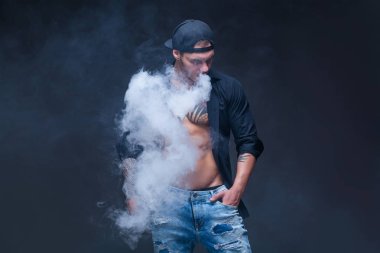 Vaper. Adam mavi kot pantolon giymiş, siyah gömlek ve siyah beyzbol şapkası dövmeler ile koyu arka plan üzerinde elektronik sigara duman