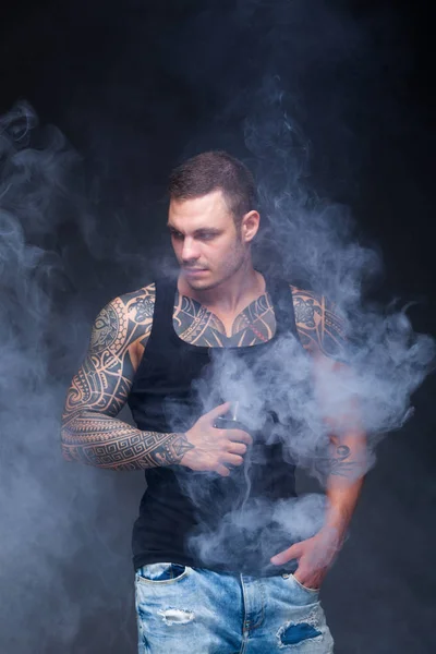 Vaper. El hombre con un torso muscular con tatuajes fuma un cigarrillo electrónico en el fondo oscuro — Foto de Stock