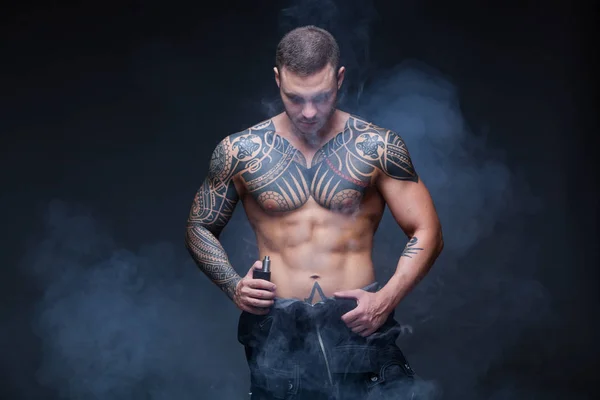 Vaper. El hombre con un torso desnudo muscular con tatuajes fuma un cigarrillo electrónico en el fondo oscuro — Foto de Stock