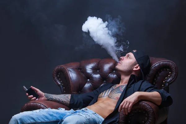 Vaper. The man with tattoos sits on a leather sofa smoke an electronic cigarette on the dark background. — Stock Photo, Image