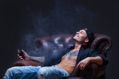 Vaper. Dövmeli adam koyu arka plan üzerinde elektronik sigara duman deri kanepede oturuyor.
