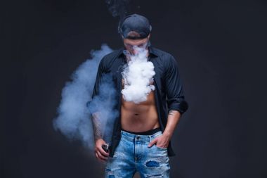 Vaper. Adam mavi kot pantolon giymiş, siyah gömlek ve siyah beyzbol şapkası dövmeler ile koyu arka plan üzerinde elektronik sigara duman