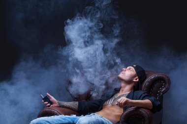 Vaper. Dövmeli adam koyu arka plan üzerinde elektronik sigara duman deri kanepede oturuyor.