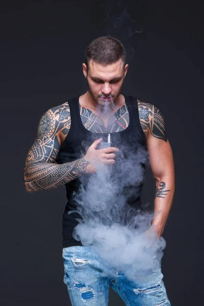 O Vaper. O homem com um tronco muscular com tatuagens fuma um cigarro eletrônico no fundo escuro — Fotografia de Stock