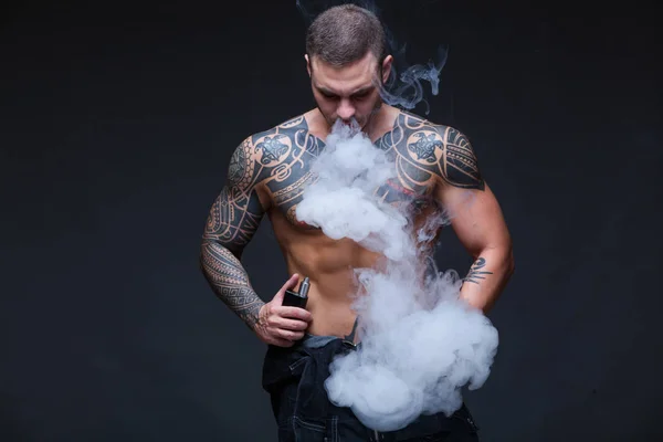 Vaper. El hombre con un torso desnudo muscular con tatuajes fuma un cigarrillo electrónico en el fondo oscuro — Foto de Stock