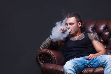 Vaper. Dövmeli adam koyu arka plan üzerinde elektronik sigara duman deri kanepede oturuyor.