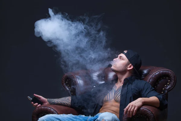 Vaper. Mannen med tatueringar som sitter på en skinnsoffa som rök en elektronisk cigarett på den mörka bakgrunden. — Stockfoto
